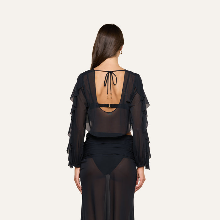 Wearing the Ambala Mesh Cover-Up Top, a woman stands facing away; the sheer black top features layered ruffle sleeves and an open back with a tie. She completes her effortlessly chic beachside style with a sheer black skirt over dark undergarments.