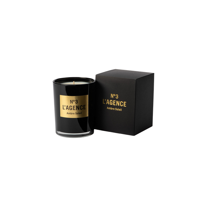 A black Ambre Soleil candle, crafted from soy-blend wax and adorned with a gold accent, sits beside its matching black and gold box on a plain white background.