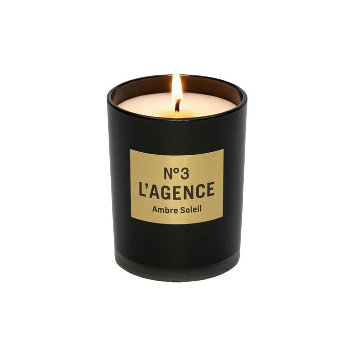A lit Ambre Soleil candle in a black glass holder, made from soy-blend wax, is displayed on a plain white background.