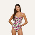 A woman stands against a plain light background, looking at the camera with her hands on her hips, wearing the Amie Bandeau One-Piece Swimsuit—a chic white one-piece featuring a pink floral and leopard print design that combines style and comfort.