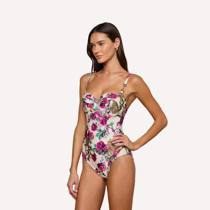 A woman stands with one hand on her hip against a plain background, wearing the Amie Bandeau One-Piece Swimsuit featuring UPF 50 protection, pink and green floral patterns, and gold accents.