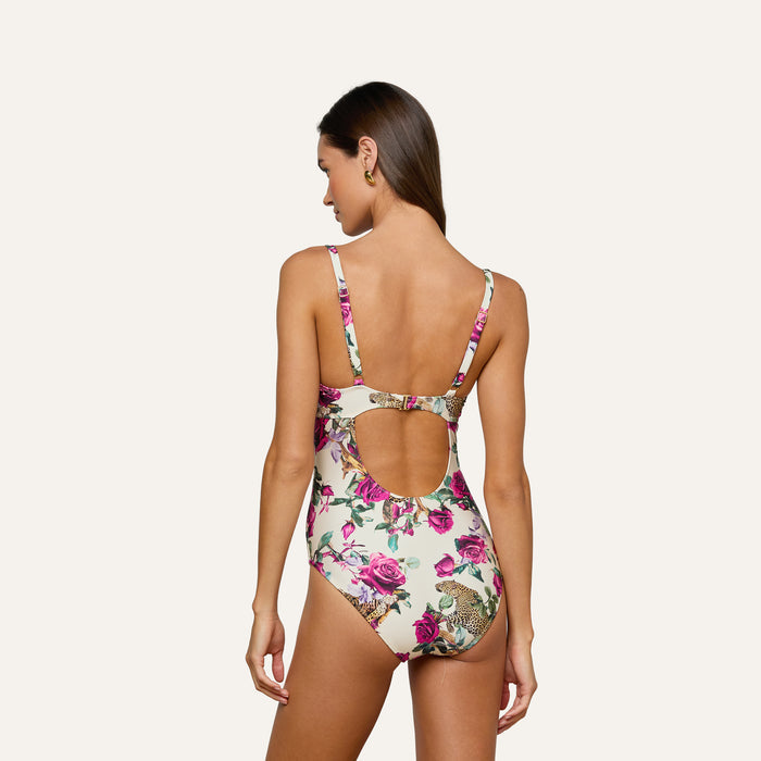 A woman faces away, wearing the Amie Bandeau One-Piece Swimsuit with a leopard print and pink floral accents. This swimsuit features a low scoop back with a circular cutout and provides UPF 50 sun protection. The background is plain and light-colored.