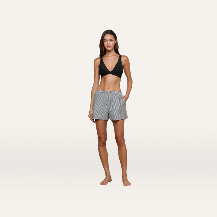 A woman stands barefoot against a plain white background, wearing a black bralette and the Amiya Cover-Up Short in tailored cotton poplin with gray stripes, her hands casually in the pockets.