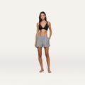 A woman stands barefoot against a plain white background, wearing a black bralette and the Amiya Cover-Up Short in tailored cotton poplin with gray stripes, her hands casually in the pockets.