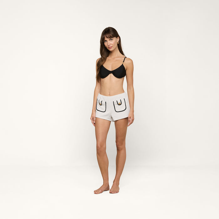 A woman stands barefoot against a plain background, wearing a black bra and the Amiya Cover-Up Short in light gray terrycloth, featuring two outlined pockets and black detailing.