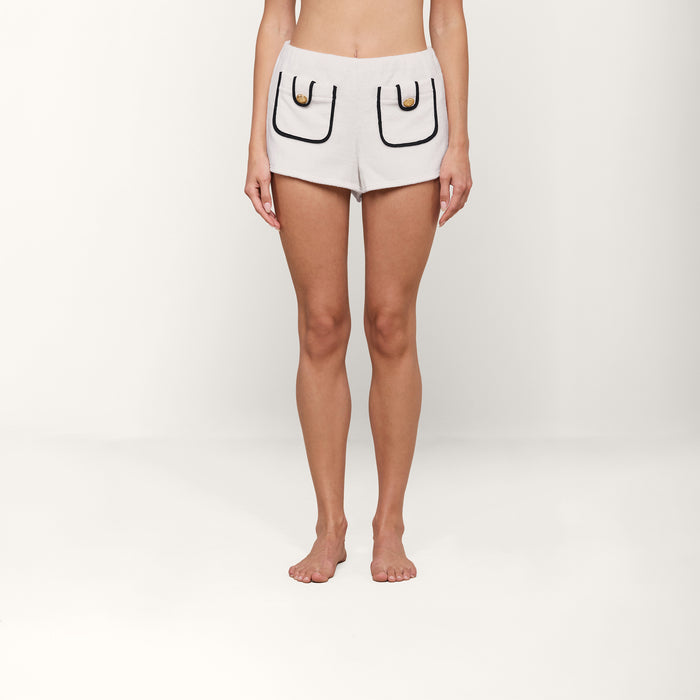 A person stands barefoot against a plain white background, wearing the Amiya Cover-Up Short—white terrycloth shorts featuring large front pockets outlined in black with gold accents—perfect for poolside lounging.