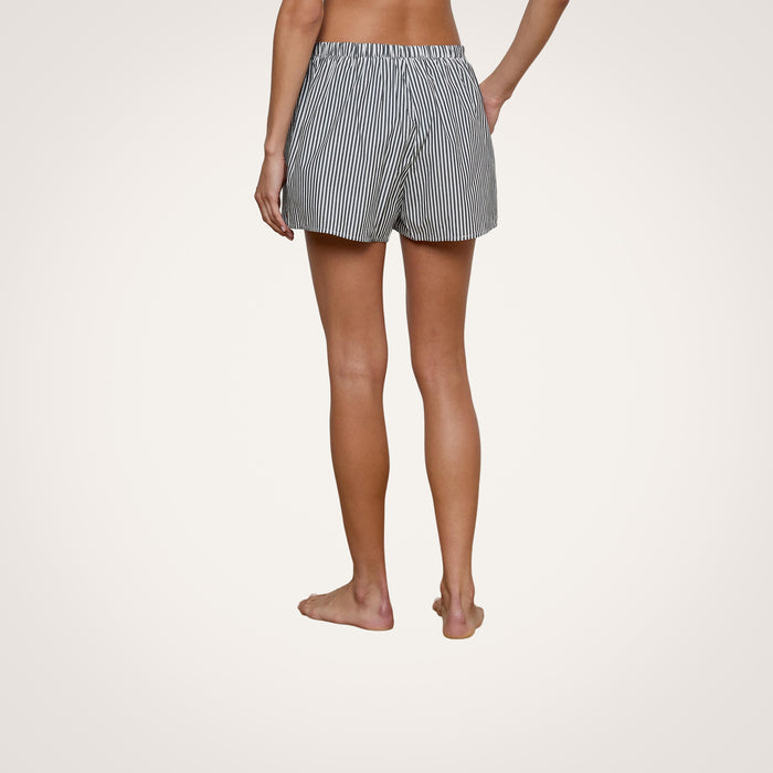 A person standing barefoot on a plain background, shown from the back, wearing the Amiya Cover-Up Short—tailored black and white vertical striped cotton poplin shorts with an elastic waistband, perfect for resort dressing.