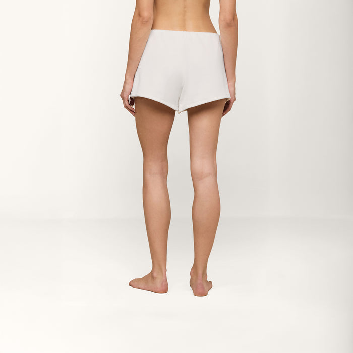 A person is shown from behind, barefoot on a plain white surface, wearing the Amiya Cover-Up Short in a light terrycloth fabric and no top. The simple, neutral background evokes relaxed poolside lounging.