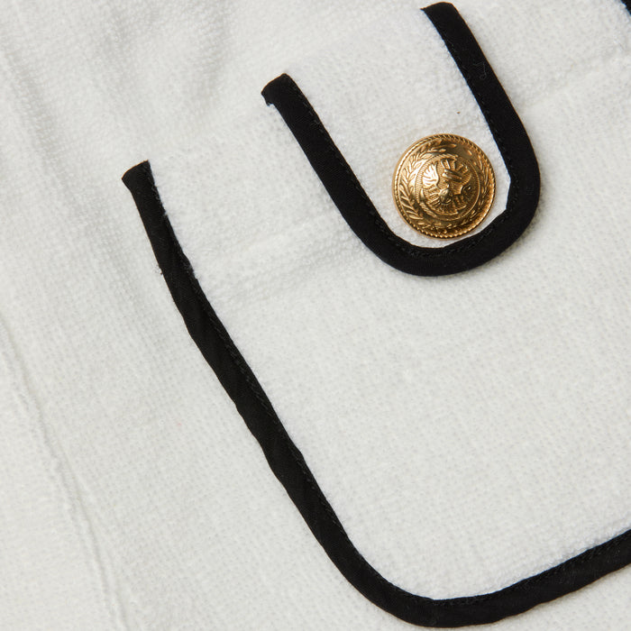Close-up of a white textured Amiya Cover-Up Short pocket with black trim and a decorative gold button featuring an embossed design, reminiscent of classic terrycloth shorts perfect for poolside lounging.
