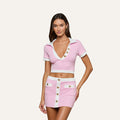 A woman wearing the Anastasia Cropped Rashguard set in light pink—a two-piece ensemble featuring a cropped, short-sleeve polo top and matching mini skirt with white trim and oversized gold buttons, all crafted from water-safe bouclé fabric—stands against a plain background.