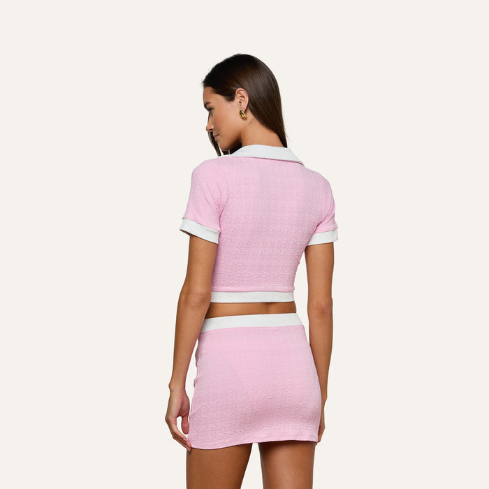 Here’s a rewrite using the provided product name: A woman stands facing away, wearing the Anastasia Cropped Rashguard—a pink textured polo top with white trim—paired with a matching pink mini skirt crafted from water-safe bouclé fabric, set against a plain light background.