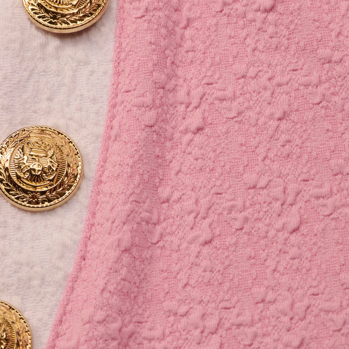 Four ornate gold buttons with engraved details are sewn onto light pink, water-safe bouclé fabric, arranged vertically along the edge of the Anastasia Cropped Rashguard.