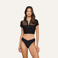 Woman wearing the Anastasia Swim Top—a black short-sleeve crop top with button details and a mesh panel—paired with high-waisted black bikini bottoms, exuding beachside sophistication against a plain background.