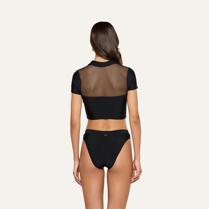 A woman stands with her back to the camera, showcasing the Anastasia Swim Top—a black short-sleeve mesh crop top—paired with black bikini bottoms for a look of beachside sophistication against a plain light background.