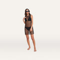 A woman wearing sunglasses, a black bikini, and the Angela Mesh Cover-Up Dress with black crystal embellishments stands barefoot against a plain, light background.
