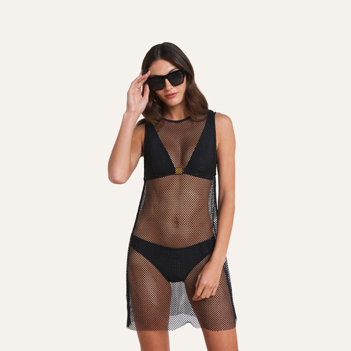 A woman wearing the Angela Mesh Cover-Up Dress— a black mesh minidress with black crystal embellishments layered over a black bikini— stands against a plain light background.