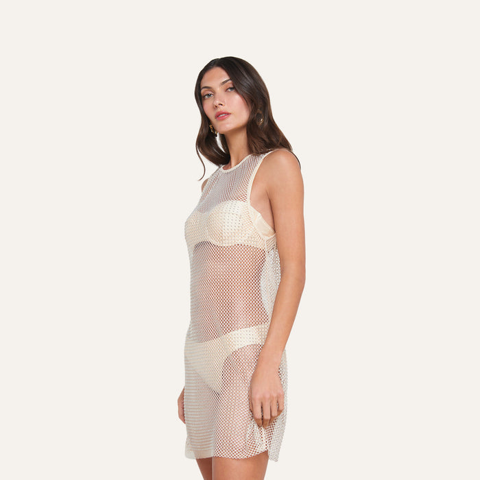 A woman stands against a plain background, modeling the Angela Mesh Cover-Up Dress as a chic swimsuit coverup over light-colored undergarments. She has long, wavy brown hair and a neutral expression, with one arm relaxed by her side.