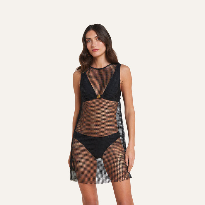 A woman stands against a plain light background wearing the Angela Mesh Cover-Up Dress—a sleeveless, see-through mesh minidress swimsuit coverup adorned with black crystal embellishments—over a black bikini.