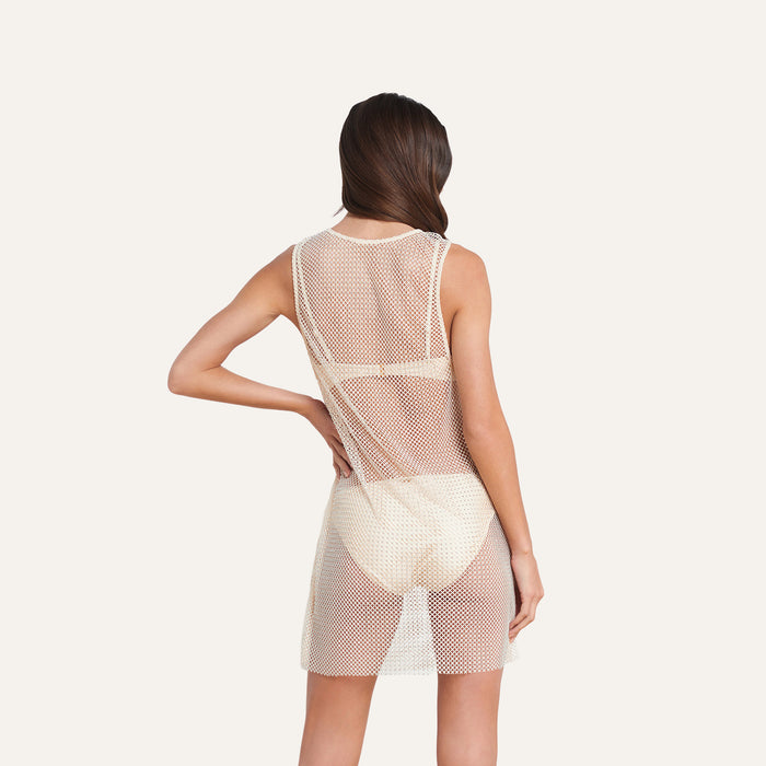 A woman stands with her back to the camera, wearing the sleeveless Angela Mesh Cover-Up Dress in white over light-colored underwear. Her right hand rests on her hip, and her long hair is parted to the side. The plain, light background highlights how this mesh dress serves as a chic and stylish swimsuit cover-up.