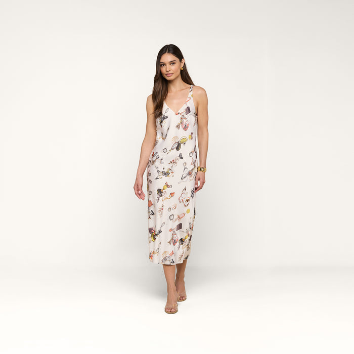 A woman stands against a plain white background wearing the Anika Slip Dress—a sleeveless, bias-cut satin white midi dress with a floral print—paired with strappy sandals.