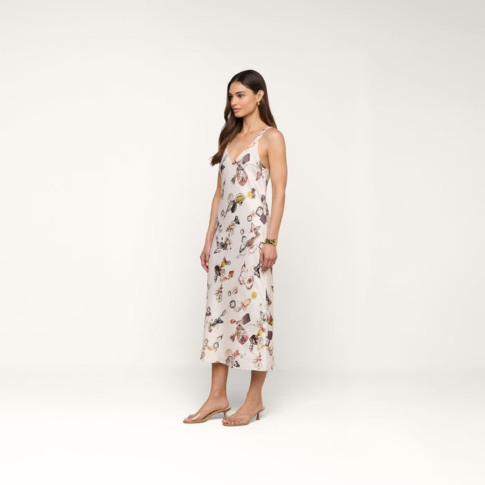 A woman stands in profile against a plain white background, wearing the Anika Slip Dress—a sleeveless, light-colored, ankle-length satin dress cut on the bias with a floral pattern—paired with sandals.