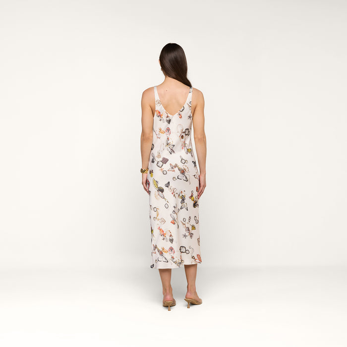 A woman with long brown hair stands facing away, wearing the Anika Slip Dress—a bias-cut satin slip dress featuring a floral pattern and a mid-length hem. She is also wearing heeled sandals, and the background is plain white.