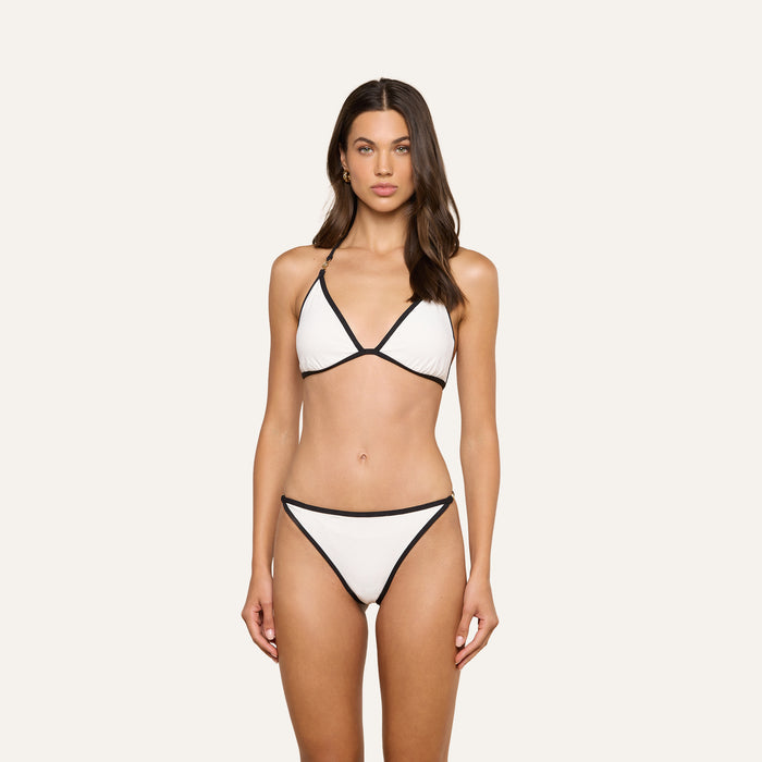 A woman stands facing forward, wearing the Annabelle Bikini Top—a white triangle silhouette bikini top with black trim and an adjustable fit. She has long brown hair and a neutral expression, posed against a plain light background.