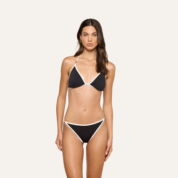 A woman stands facing forward against a plain light background, wearing the Annabelle Bikini Top—a black triangle bikini with white trim and an adjustable fit.