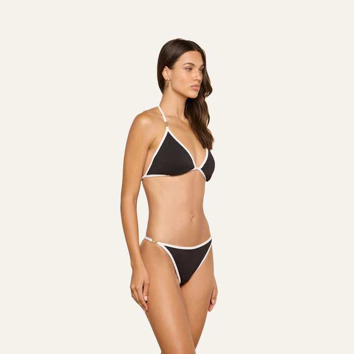 A woman stands against a light background, wearing the Annabelle Bikini Top, a black triangle bikini with white trim and an adjustable fit for comfort. She is looking slightly to her left.