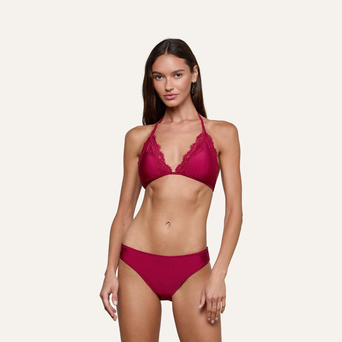 A woman stands against a plain background, wearing the Annabelle Triangle Bikini Top in deep red, featuring adjustable ties and lace trim. She has long brown hair and gazes at the camera with a relaxed expression.