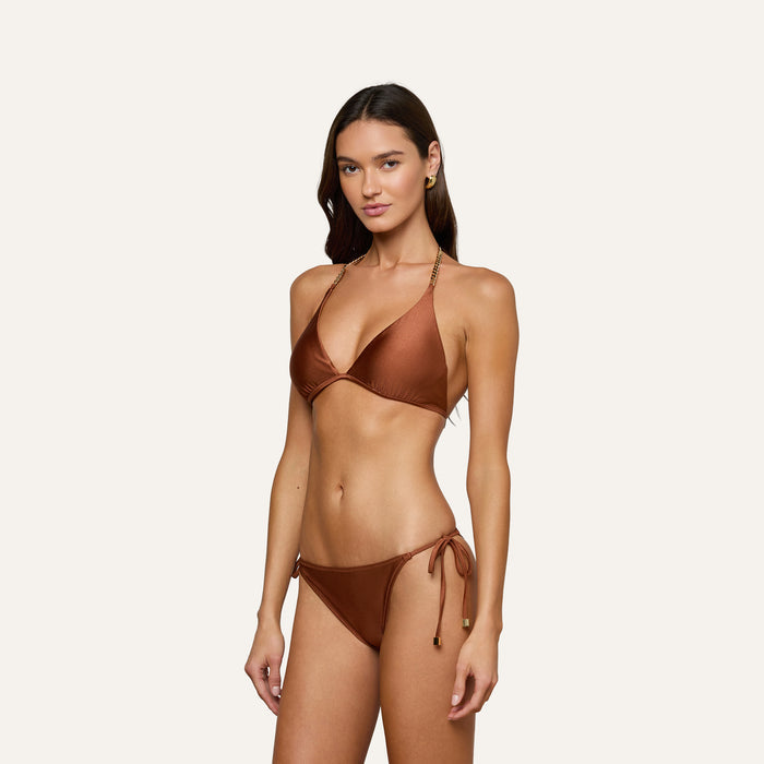 A woman poses against a plain light background wearing the Annabelle Triangle Bikini Top in bronze, featuring luxe hardware, a halter neck design, and adjustable tie-side bottoms for a customizable fit.