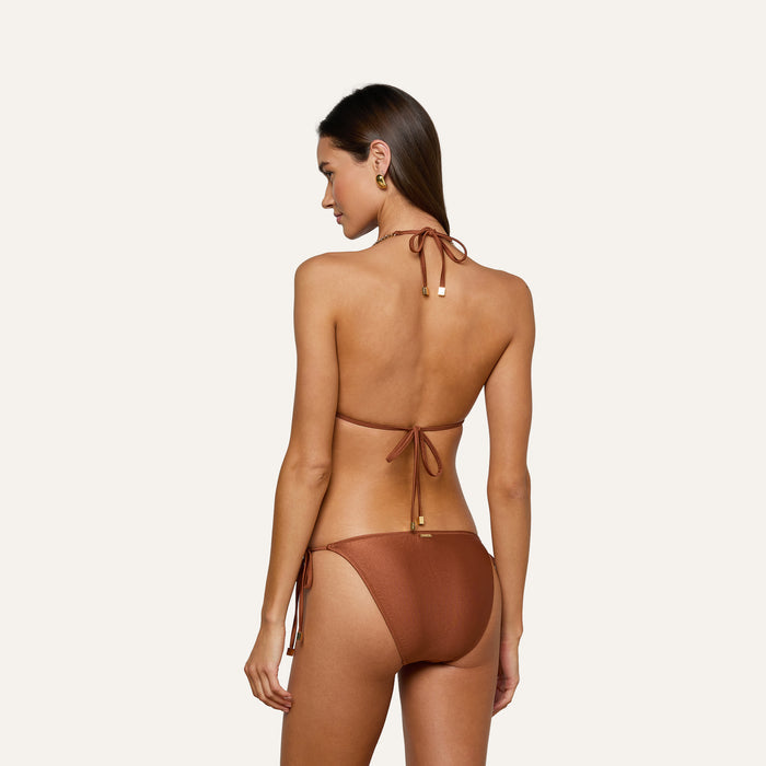 A woman stands facing away against a plain light background, wearing the Annabelle Triangle Bikini Top in bronze, featuring a tie-back and tie-side design for an adjustable fit.