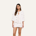 A woman stands against a plain light background wearing the Anouk Jean Jacket over a matching white mini dress. She has long brown hair and looks forward with a neutral expression.