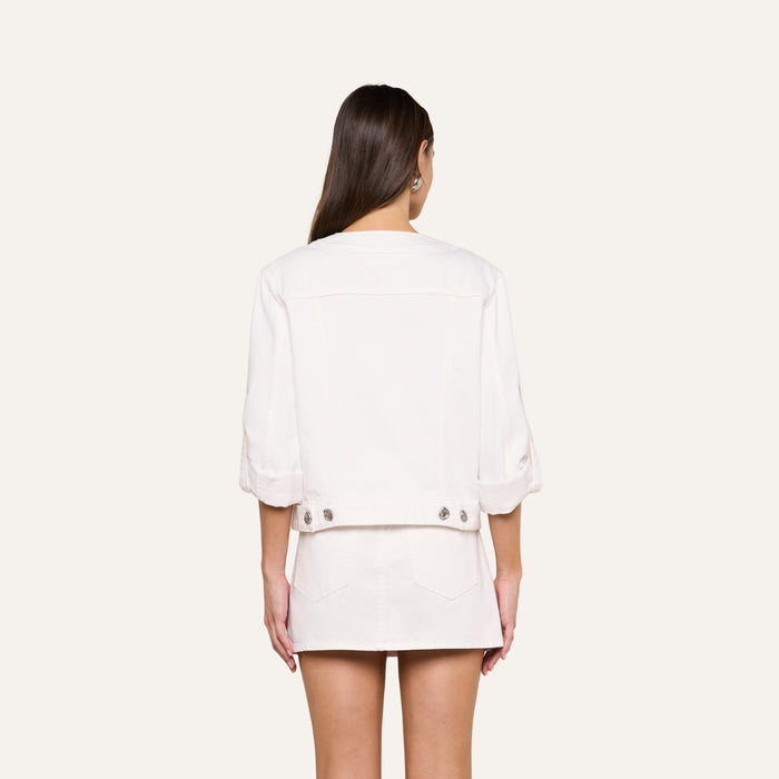 A woman stands facing away, wearing the Anouk Jean Jacket with rolled sleeves and a matching skirt. Her long brown hair is down, and her hands rest by her sides against a plain background.