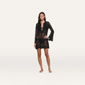 Barefoot woman wearing the Ansley Cover-Up Dress—a black, long-sleeved, sheer lace romper with a deep V neckline—styled as a chic boho coverup against a plain white background.