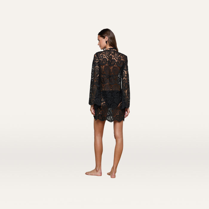 A woman stands barefoot with her back to the camera, wearing the Ansley Cover-Up Dress—a black, long-sleeve paisley crochet design that is semi-transparent. The background is plain and light-colored.
