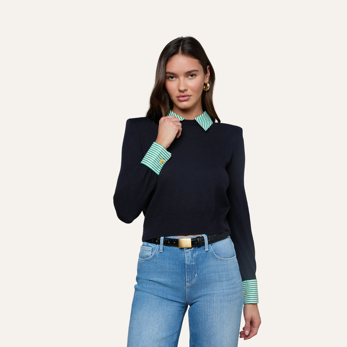 A woman stands against a plain light background wearing the April Poplin Collar Pullover—navy with green striped cuffs and a striped poplin collar—paired with light blue jeans and a black belt.