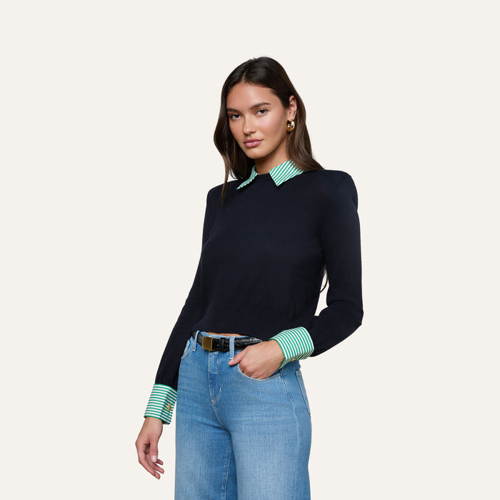 Wearing the April Poplin Collar Pullover layered over a green and white striped shirt, a woman pairs it with light blue jeans and a black belt, standing against a plain light background.