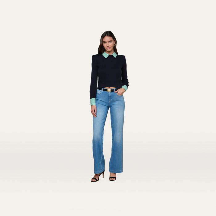 A woman stands against a plain background wearing the April Poplin Collar Pullover styled over a light green striped shirt, paired with blue jeans and black heeled sandals. She has long dark hair and poses with a relaxed, confident expression.