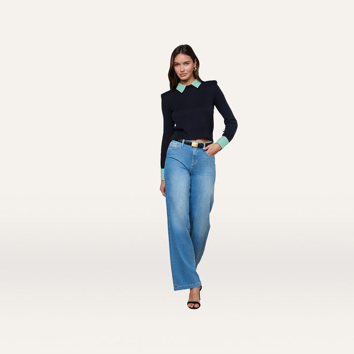 A woman poses against a plain background, wearing the April Poplin Collar Pullover—a black sweater with mint green cuffs and a striped poplin collar—paired with blue wide-leg jeans, a black belt, and black heels.