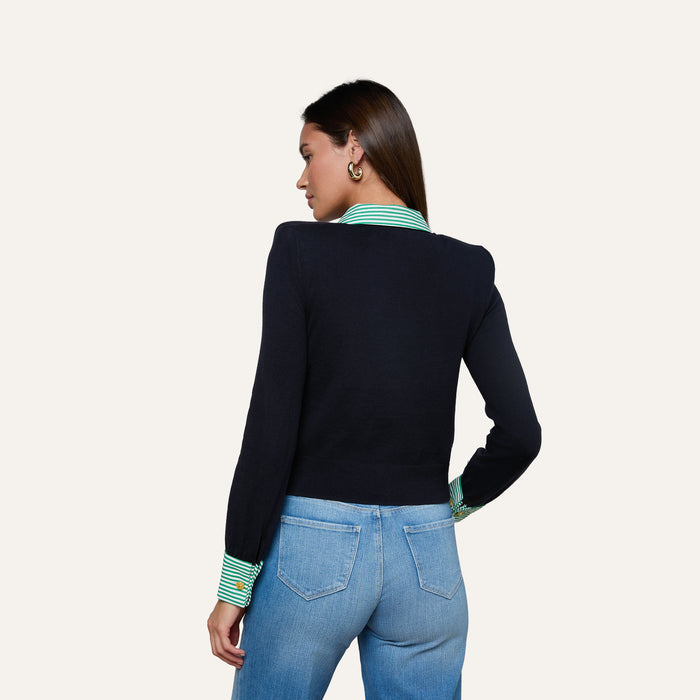 A woman faces away, wearing the April Poplin Collar Pullover—a black sweater featuring a striped poplin collar and green striped cuffs—paired with light blue jeans, set against a plain, light-colored background.