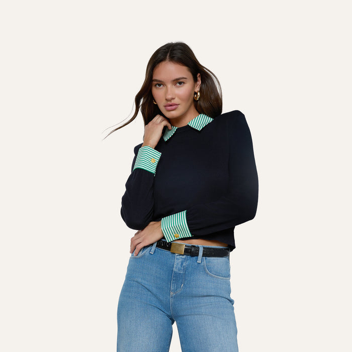 A woman models the April Poplin Collar Pullover, featuring a navy design with a green striped poplin collar and cuffs. She pairs it with blue jeans and a black belt, posing with one hand on her chin and the other resting on her hip against a plain background.