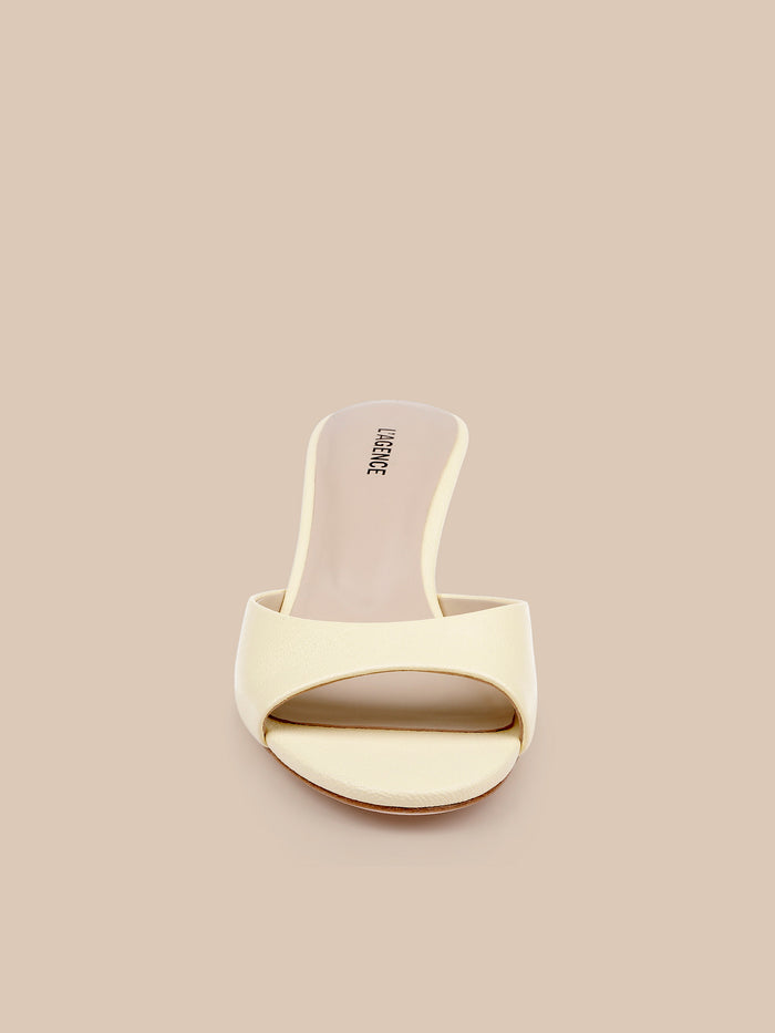 The Arleen Kitten-Heel Mule is a cream-colored leather open-toe sandal featuring a wide strap across the front, showcased against a beige background in a front-facing view.