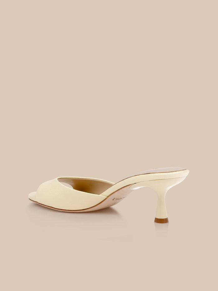 The Arleen Kitten-Heel Mule, in cream-colored leather with an open toe and a low, curved heel, is displayed against a matching beige background from a rear side angle.