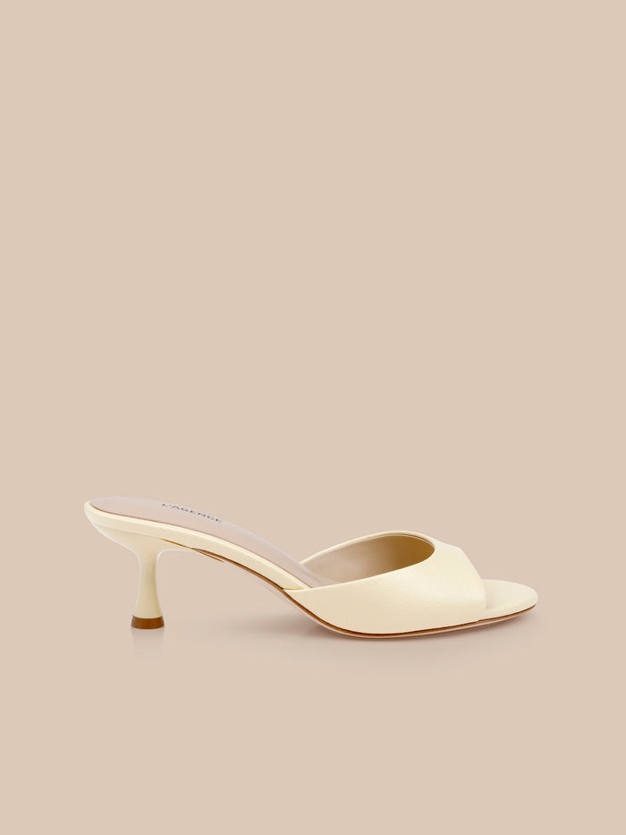The Arleen Kitten-Heel Mule, a single cream-colored leather sandal featuring an open toe and a low, curved kitten heel, is displayed on a solid beige background.