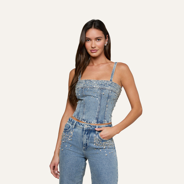 Wearing the Arlin Embellished Tank, a woman stands against a plain light background, pairing the crystal-studded, cropped denim top—adorned with pearls and studs—with matching jeans.