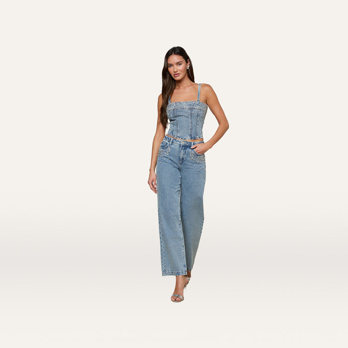 A woman stands against a plain background wearing the Arlin Embellished Tank—a cropped light blue denim corset top—paired with high-waisted wide-leg jeans featuring crystal-studded pocket details and open-toed heels.