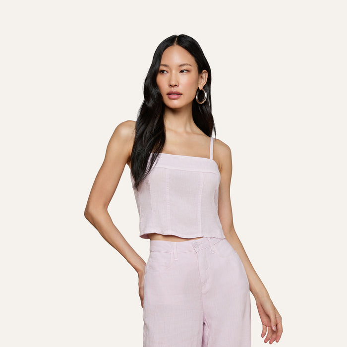 Wearing the Arlin Linen Tank, a woman with long black hair poses with one hand on her hip against a plain background, pairing the light pink cropped corset top with matching high-waisted pants.