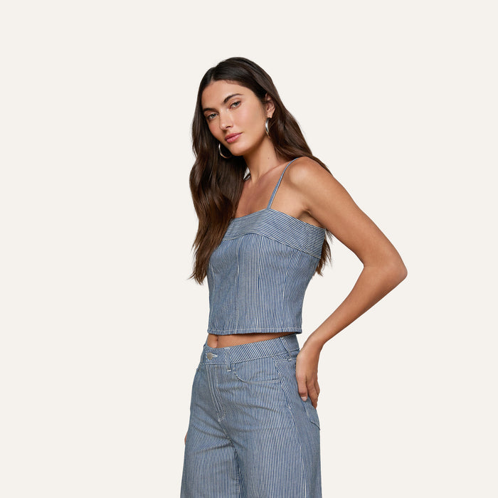 Woman wearing the Arlin Tank, a blue stripe sleeveless corset top paired with matching high-waisted pants, standing against a plain light background and looking at the camera with one hand on her hip.