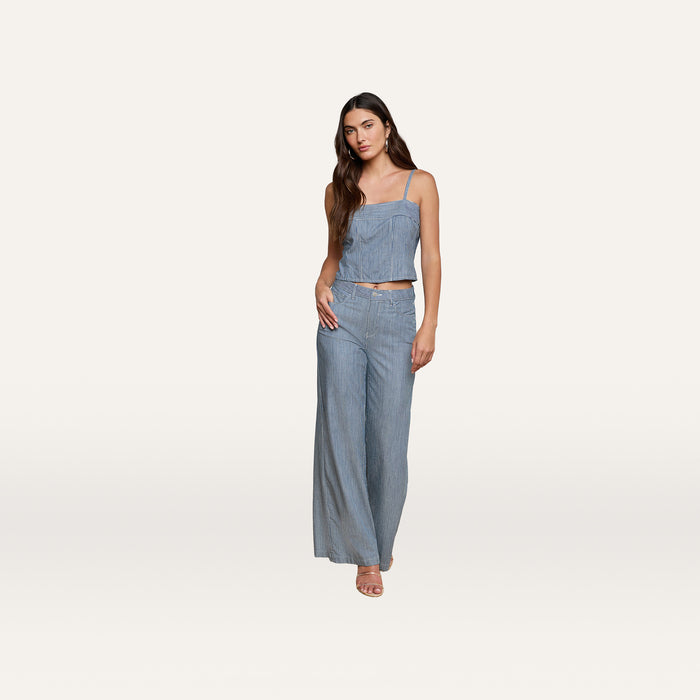 A woman wearing the Arlin Tank— a sleeveless denim corset top— paired with matching wide-leg pants, stands against a plain white background with one hand on her hip and the other relaxed by her side.